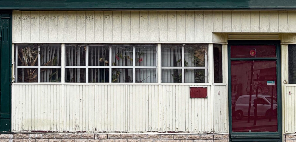 Façade d’un bâtiment à revêtement clair avec soubassement en pierre.
Une large fenêtre à montants blancs occupe la partie gauche, laissant voir des plantes derrière des stores verticaux.
À droite, une porte vitrée rouge encadrée de vert porte le numéro 4.