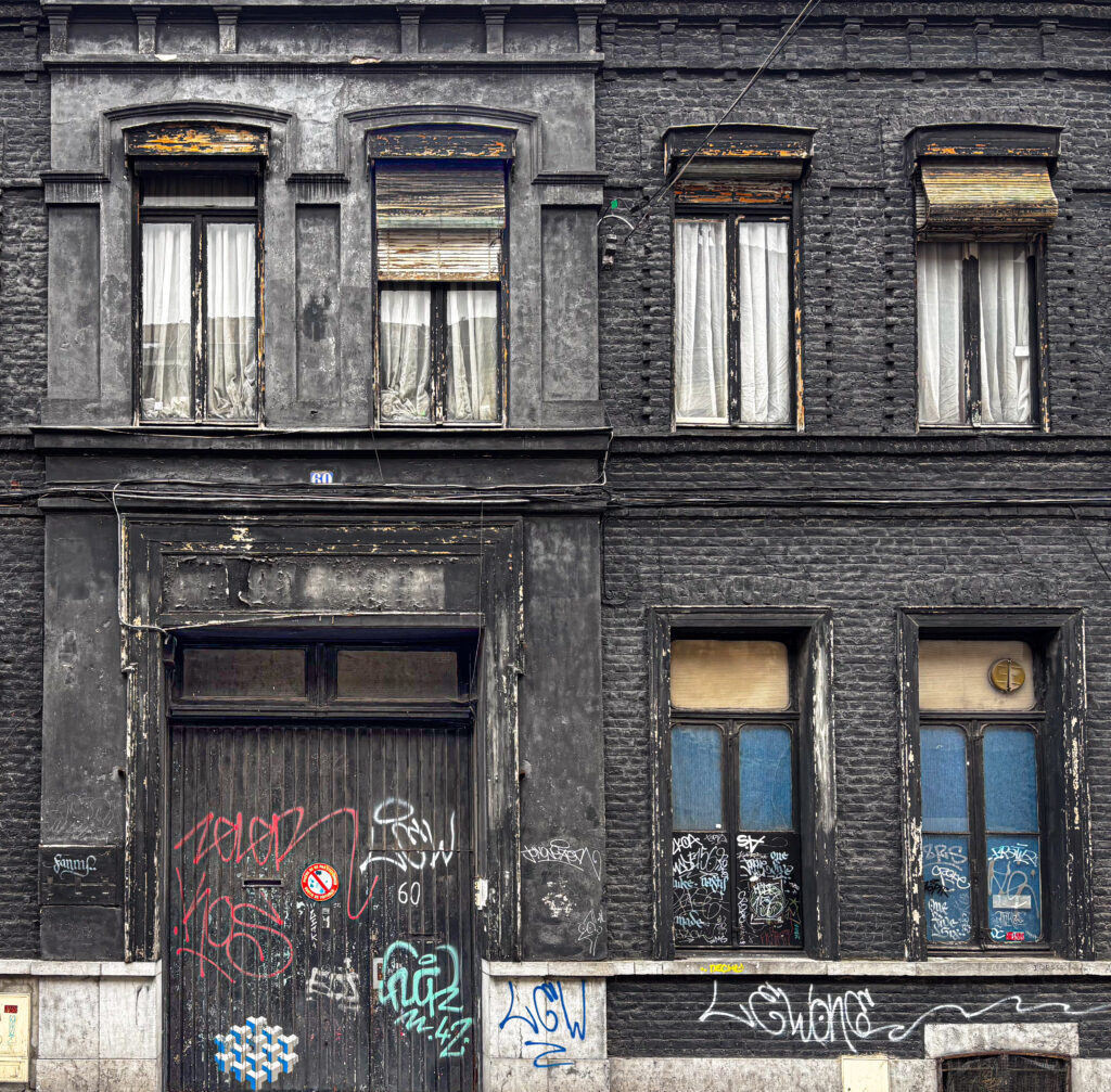 Façade d’un immeuble ancien en briques peintes en noir, à deux niveaux.
En bas, une grande porte en bois et deux fenêtres, tous recouverts de graffitis et marqués par l’usure.
En haut, quatre fenêtres étroites avec rideaux ou volets, encadrements dégradés et peinture écaillée.